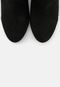 Bottes noires en daim à la cheville avec un bout pointu, une texture lisse et sans quincaillerie ni motifs visibles, affichant un design épuré et minimaliste.