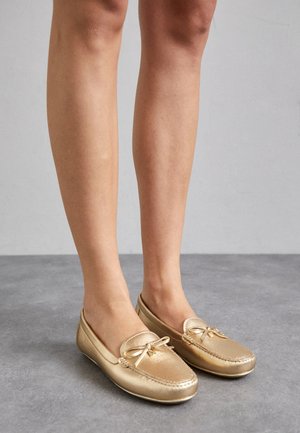 Fødder iført metalliske guld loafers med sløjfedetalje, stående på en grå gulv mod en ensfarvet grå baggrund.