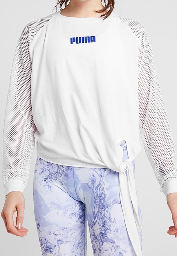 Donna che indossa una maglietta bianca a maniche lunghe PUMA con maniche in rete e leggings a motivo marmo lilla, che sta facendo un nodo sul lato della maglietta.