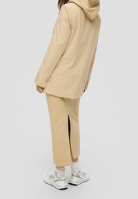 Osoba v béžovém pruhovaném saku s kapucí, béžové midi sukně s rozparkem vzadu, bílých ponožkách a bílých teniskách, stojící na šedém pozadí.