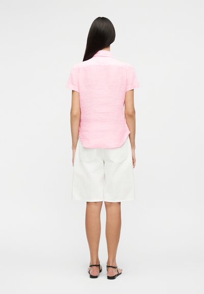 Femeie stând cu spatele la cameră, purtând o bluză roz cu mâneci scurte, pantaloni scurți albi până la genunchi și sandale negre, pe un fundal simplu.