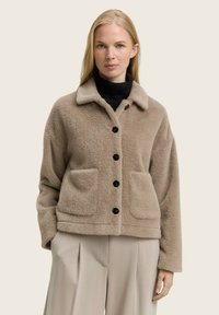 Beige, harige, kraagloze jas met twee voorkeurszakken en vijf zwarte knopen in het midden. Zachte textuur, ontspannen pasvorm.