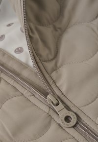 Beige quiltad jacka med en slät yta, som har dragkedjestängning och ett mönstrat innerfoder med smiley-design.
