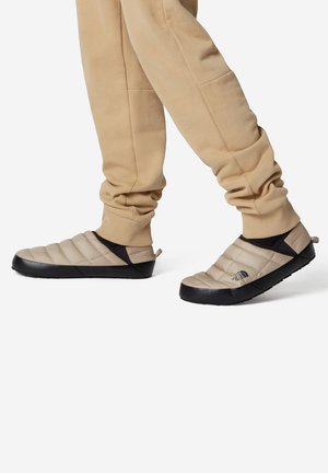 Zapatos tipo slip-on acolchados en color beige con una suela de goma negra y un upper texturizado, que presenta un patrón de acolchado diagonal y acentos elásticos.