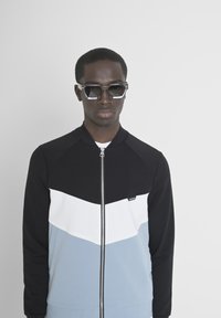 Chaqueta con cremallera en negro, blanco y azul claro con un patrón de chevrón; presenta un cuello acanalado y mangas largas. El modelo lleva gafas de sol angulares.