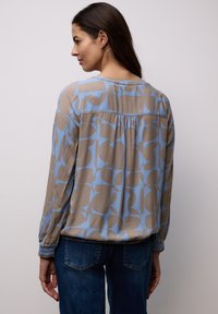 Femme de dos portant un chemisier à manches longues avec des motifs géométriques abstraits bleus et beiges, et un jean bleu foncé.