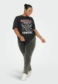 Kobieta uśmiechająca się, ubrana w czarną koszulkę "Rock & Boots Cowgirl", szare dżinsy w stylu acid-wash oraz białe trampki, stojąca na białym tle.