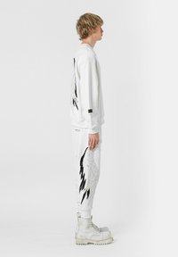 Sudadera blanca y pantalones con patrones de alas en negro y plata. Modelo usando botas blancas gruesas. Diseño minimalista con detalles bordados.