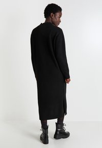 Robe-pull longue noire avec des détails côtelés, fente sur le côté, texture douce, portée avec des bottes à plateforme noires à lacets et des chaussettes sombres.