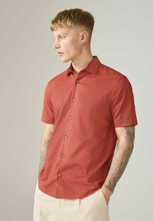 Young man with short hair and arm tattoos wearing a rust-colored short-sleeve button-up shirt and light beige pants, hands in pockets.