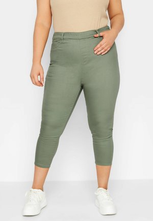 Dorosła osoba ubrana w oliwkowozielone skrócone spodnie i białe trampki, stoi z jedną ręką na biodrze, a drugą swobodnie opuszczoną wzdłuż ciała.