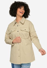 Giacca camicia in denim beige con bordi sfilacciati, due tasche sul petto e chiusura a bottoni. Presenta un colletto classico e maniche lunghe.