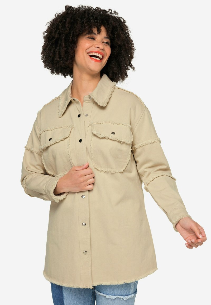 Beige denimskjorta med fransade kanter, två bröstfickor och knappstängning. Har en klassisk krage och långa ärmar.