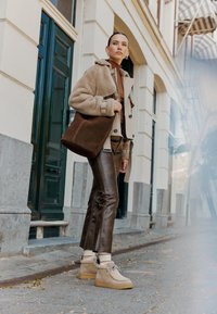 Manteau en shearling marron sur un haut beige, pantalon en cuir marron ajusté et bottes en suede beige avec des détails texturés, accompagnés d'un sac à épaule marron.