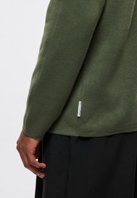 Πράσινο πουλόβερ ελιάς, υφή πλεκτή, στενά μακριά μανίκια, λεπτομέρεια πλευρικής σχισμής και μικρή ετικέτα λογότυπου στο τελείωμα. Φορεμένο πάνω από σκούρο παντελόνι.