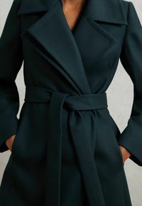 Naine kannab tume roheline vööga mantel, millel on laiad kraed ja käed taskutes, neutraalse tausta taustal.