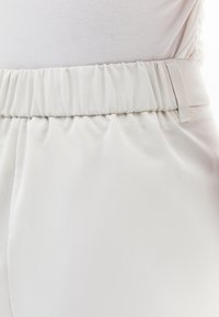 Witte elastische tailleband met geraffineerde textuur, leidend naar een gladde stof die in een ontspannen silhouet valt. Eenvoudig en schoon ontwerp.