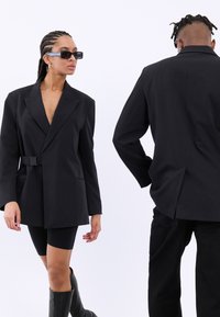 Schwarzer übergroßer Blazer mit einem Gürtel, breiten Revers und aufgesetzten Taschen. Gestylt mit schwarzen Shorts und kniehohen Stiefeln, die eine glatte Textur haben.