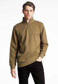 Felpa verde oliva con zip a quarto, polsini a coste e logo decorativo sul petto. Tessuto morbido, vestibilità comoda, abbinata a pantaloni neri.