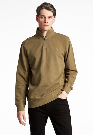 Olive green sweatshirt with a quarter zip, ribbed cuffs, and a decorative logo on the chest. Smooth texture, relaxed fit, styled with black pants.
