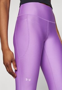 Lila atletiska leggings har en mjuk textur, hög midja och en sidficka. Designen inkluderar en liten vit logotyp nära fållen.