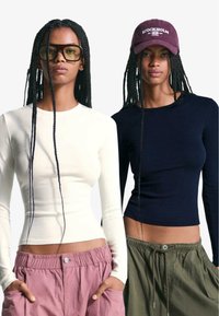 Două bluze cu mâneci lungi, una albă și una bleumarin, asortate cu pantaloni roz și verzi, cu croială lejeră, evidențiind o textură fină a materialului.