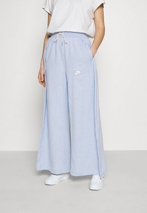 Person iført lyseblå Nike sweatpants med vide ben, hvid t-shirt og hvide sneakers, stående mod en ensfarvet lys baggrund.