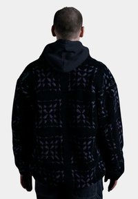 Chaqueta de forro polar negra con un patrón geométrico en púrpura y azul, que cuenta con una capucha y un logo verde en el hombro izquierdo.