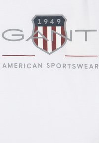 Hvit t-skjorte med "GANT" logo i grå, med skjolddesign og røde og hvite striper over "1949" og teksten "AMERICAN SPORTSWEAR" nedenfor.