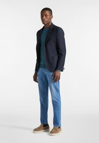 Marinblå kavaj över en tealtröja, matchad med ljusblå jeans och beigea slip-on skor. Kavajen har detaljrika fickor och ett mjukt tyg.
