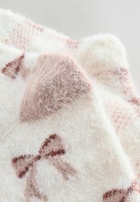 Kudottu heittotaipe pehmeässä kerma- ja vaaleanpunaisessa sävyssä, jossa on pörröinen tekstuuri, ruskeita nauhakuvioita ja hienovaraisia värieroja.