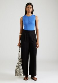 Vrouw in een blauwe mouwloze ribbed top, zwarte wijde broek, zwarte sandalen, met een tas met zebraprint, staande tegen een effen achtergrond.