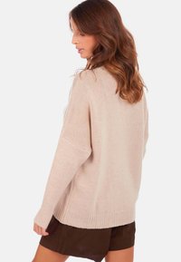 Pull en maille beige à coupe décontractée, manches larges et ourlet côtelé. La texture semble douce avec un motif subtil et délicat.