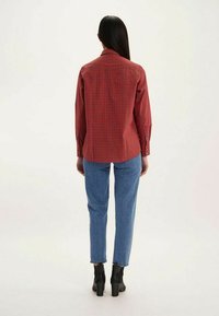 Chemise à manches longues à carreaux rouges avec une coupe droite, des poignets boutonnés et un empiècement au dos, associée à un jean bleu clair et des bottes à talons noires.