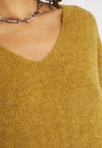 Musztardowy sweter w serek z materiału w prążki. Charakteryzuje się miękką teksturą i dopasowanym krojem. Widoczny srebrny naszyjnik.