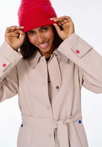 Béžový trenčkot so zapínaním na gombíky a golierom, s červenými a modrými doplnkovými gombíkmi. Modelka má na hlave jasne červenú pletenú čiapku.