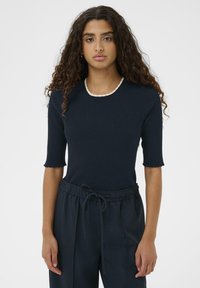 Vrouw met lang krullend haar, gekleed in een donker geribbelde top met korte mouwen en een gegolfde witte halslijn, met bijpassende donkere broek met trekkoord.