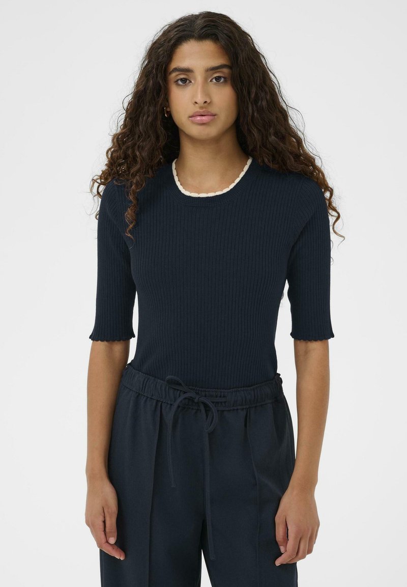 Vrouw met lang krullend haar, gekleed in een donker geribbelde top met korte mouwen en een gegolfde witte halslijn, met bijpassende donkere broek met trekkoord.