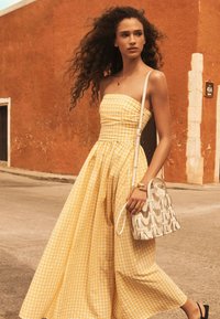 Donna con capelli ricci che indossa un vestito senza spalline a quadri giallo, tiene una borsa bianca con motivo, cammina su una strada con un muro arancione.