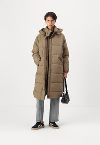 Manteau long rembourré couleur kaki avec un col montant et une capuche. Présente une fermeture éclair, des coutures visibles et des poches latérales. Porté sur un haut marron, associé à un jean clair et des baskets noires.