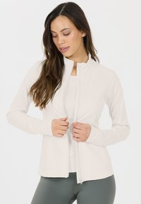 Weiße zip-up Jacke mit Stehkragen, langen Ärmeln und enganliegendem Design; aus glattem, elastischem Stoff gefertigt; über einem leichten Oberteil getragen.