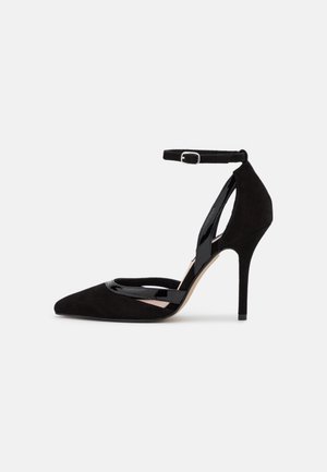 Zapato negro de tacón alto con punta afilada y tira en el tobillo, fabricado en ante y charol, con un tacón stiletto delgado.