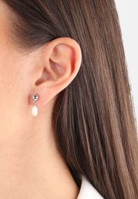 Pendiente de tono plateado con un botón redondo y un acento colgante en forma de perla. Textura suave con un acabado pulido.