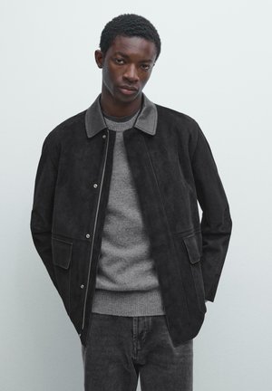 Veste en daim noir avec un col à revers, fermeture éclair, deux poches avant et une texture lisse, portée par-dessus un pull gris et un jean.