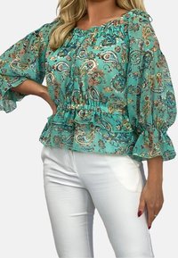 Turkos paisley-blus med långa, ballongärmar, volangkant och elastisk halsringning. Bärs med vita byxor av lätt, genomskinlig tyg.