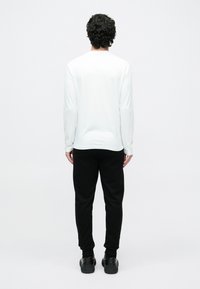 Valge pika varrukaga särk ümmarguse kaelusega, mustade kitsenevate dressipükste ja mustade tossudega, vaadates tagantpoolt.