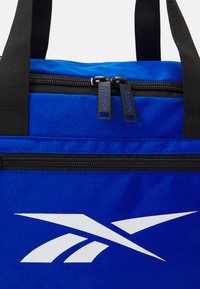 Geantă de sport din material textil albastru, cu fermoar negru, un logo Reebok alb și mânere web negre pentru o transportare ușoară.