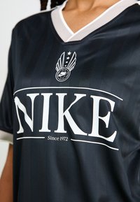 Svart Nike-drakt med V-hals, med hvit logo og teksten "NIKE" med "Siden 1972" under. Vertikal stripet teksturdesign.