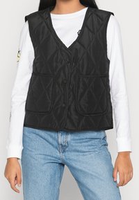 Svart vadderad väst med V-ringning, med två framfickor och tryckknappar. Bärs över en vit långärmad skjorta och blå jeans.