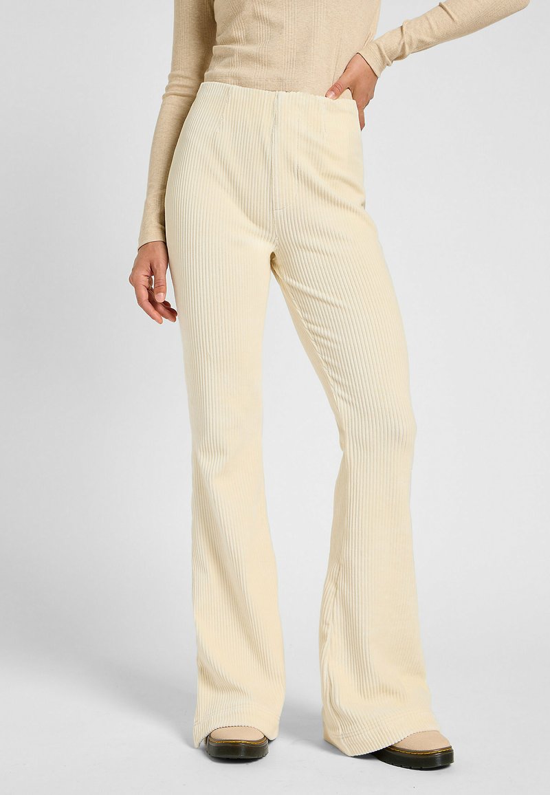 Pantaloni a zampa d'elefante in corduroy color crema con righe verticali, dotati di una vita alta e una finitura liscia. Scarpe con platform nere visibili.